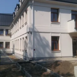 Budynek z białymi ścianami i ciemnym dachem z lukarnami, widok z zewnątrz, teren wokół w trakcie prac ziemnych.