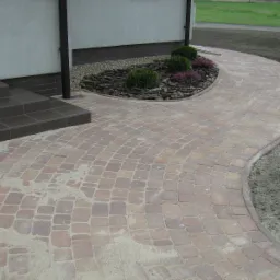 Zakrzywiony chodnik z czerwono-brązowej kostki, prowadzący od schodów wejściowych do domu, obok rabata z krzewami i korą.