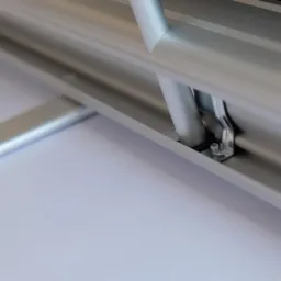 Szczegół mocowania aluminiowej konstrukcji, widoczny metalowy uchwyt z śrubą mocującą oraz fragment białej powierzchni w tle.