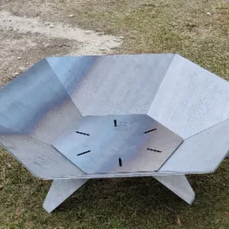 Metalowy kosz na ognisko o heksagonalnym kształcie, z widocznymi śladami spawania, ustawiony na trawie i ziemi.