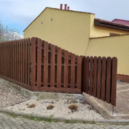 Ciemnobrązowe, drewniane ogrodzenie panelowe z furtką na betonowej podmurówce, otoczone kamieniami ozdobnymi i kwitnącym krzewem forsycji, na tle żółtego domu.