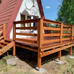 Nowo wybudowany, drewniany taras z balustradą, przylegający do domu z czerwonym dachem, widoczny z poziomu trawnika.