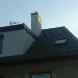 Dom z nowym dachem z blachodachówki w kolorze grafitowym, widoczny komin i okno dachowe.