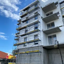 Budynek wielorodzinny w trakcie ocieplania styropianem, widoczne rusztowania z żółtymi elementami zabezpieczającymi, betonowy parter i balkony, błękitne niebo z chmurami w tle.