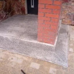 Wykończone schody wejściowe z szarego, drobnoziarnistego kamienia z czerwoną kolumną z cegły klinkierowej, prowadzące do ciemnych drzwi.