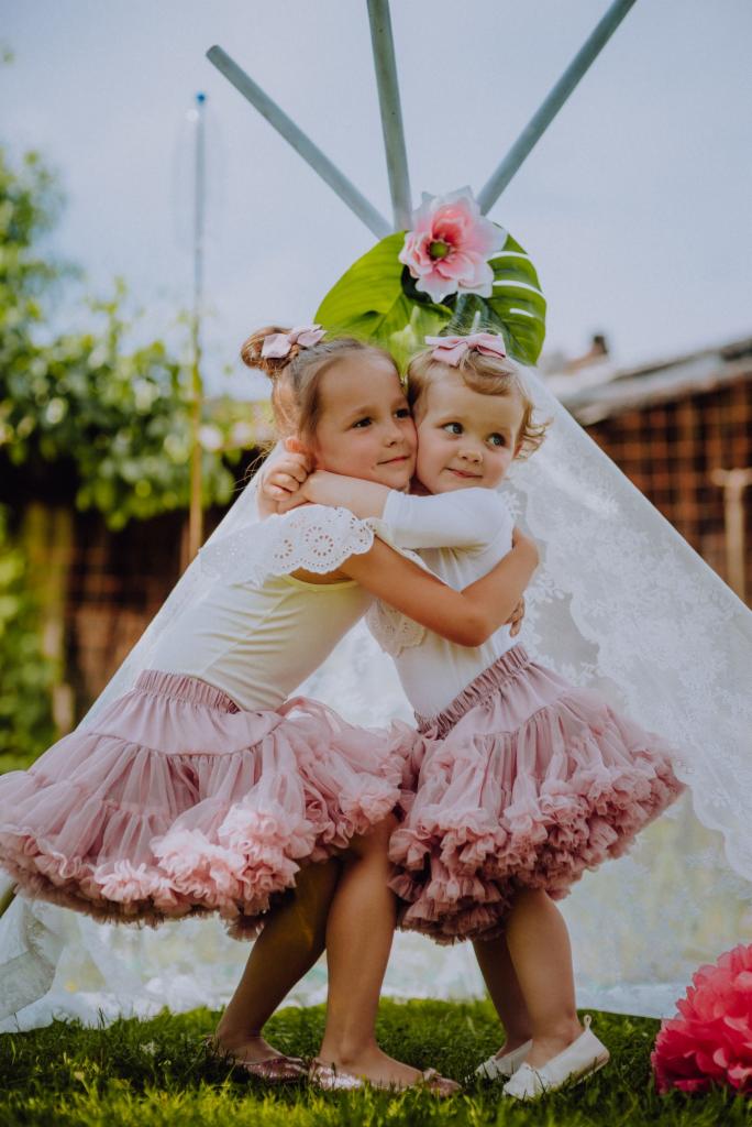 Dwie małe dziewczynki w tiulowych spódniczkach i kokardami we włosach przytulają się na tle tipi ozdobionego kwiatami i liśćmi.