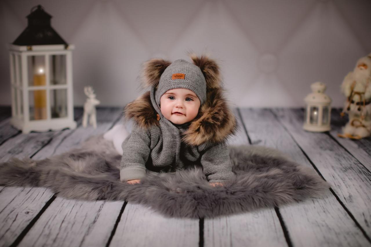 Urocze niemowlę w szarym, dzierganym kombinezonie z futrzanym kapturem, leżące na puszystym, szarym dywaniku na tle drewnianej podłogi i dekoracji świątecznych.