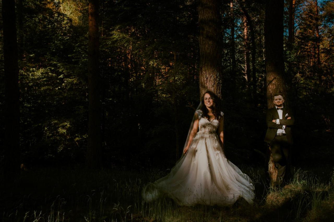 Panna młoda w koronkowej sukni ślubnej i welonie, uśmiechnięta, opiera się o drzewo w lesie; obok, w tle, pan młody w czarnym garniturze i muszce, z założonymi rękami.