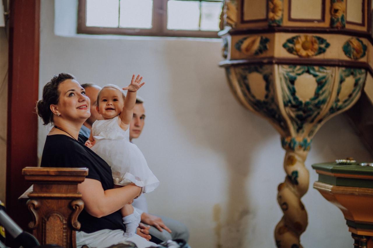 Uśmiechnięta dziewczynka w białej sukience, trzymana na rękach przez kobietę w kościele, z uniesioną rączką na tle kazalnicy.