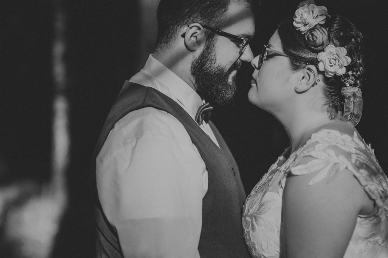 Czarno-białe zbliżenie pary w okularach, delikatnie dotykającej się nosami; on w kamizelce i muszce, ona z kwiatami we włosach i koronkową sukienką.