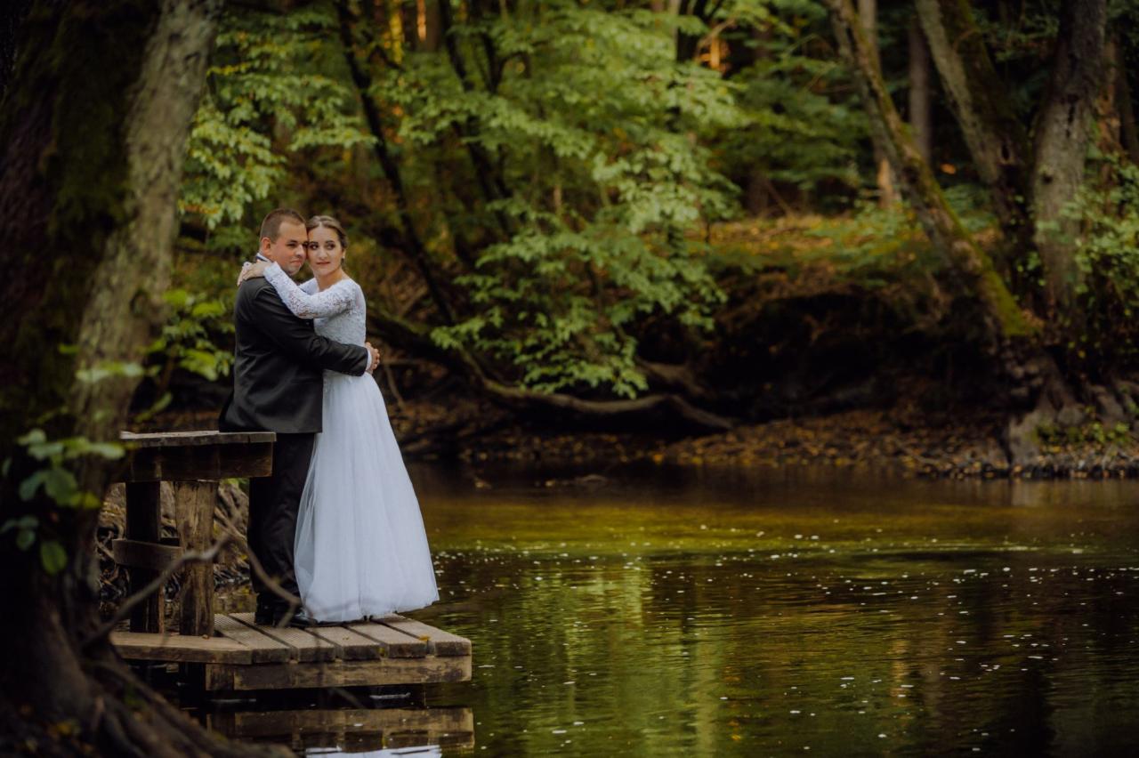 Para młoda w objęciach na drewnianym pomoście nad wodą, otoczona drzewami w leśnym otoczeniu, panna młoda w sukni z długim rękawem.
