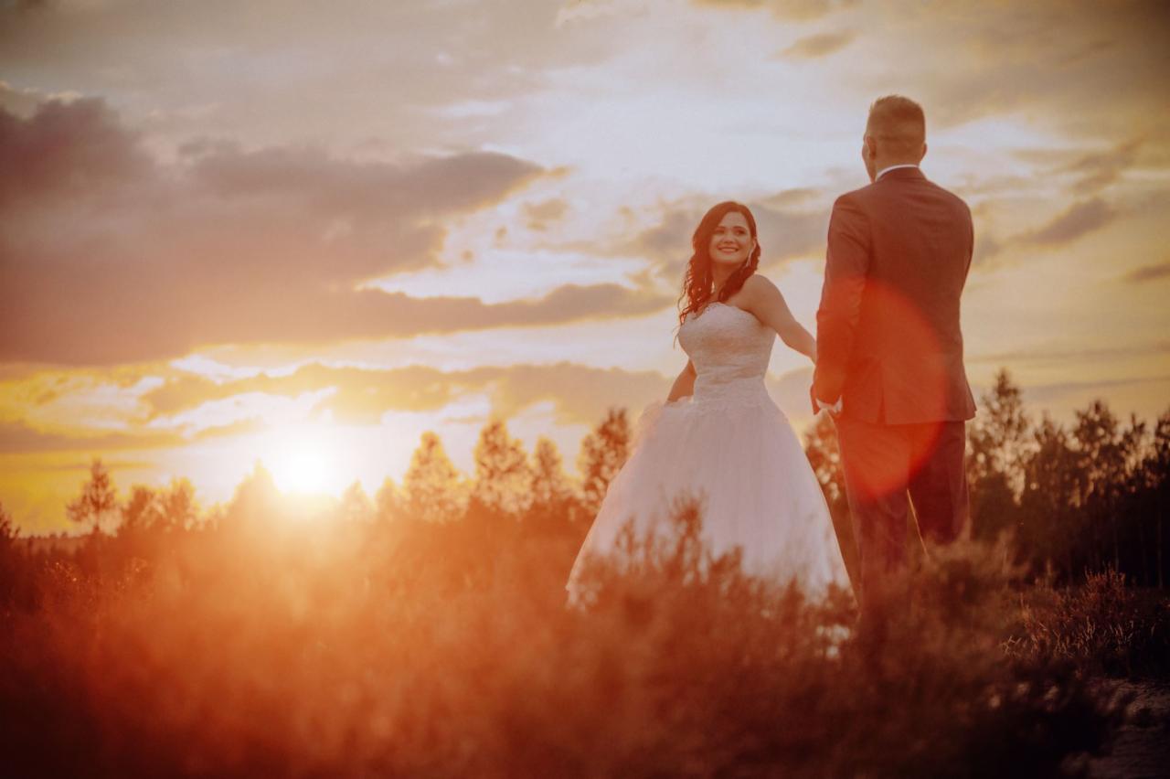 Panna młoda w białej sukni i pan młody w garniturze trzymają się za ręce na tle zachodzącego słońca i drzew, lekko rozmyte ujęcie.