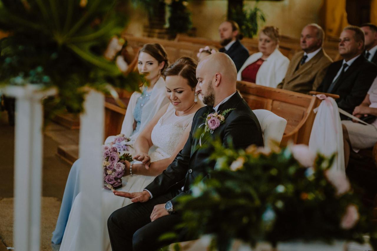 Para młoda siedzi w kościele podczas ceremonii, panna młoda trzyma bukiet fioletowych róż, w tle goście siedzący w ławkach.