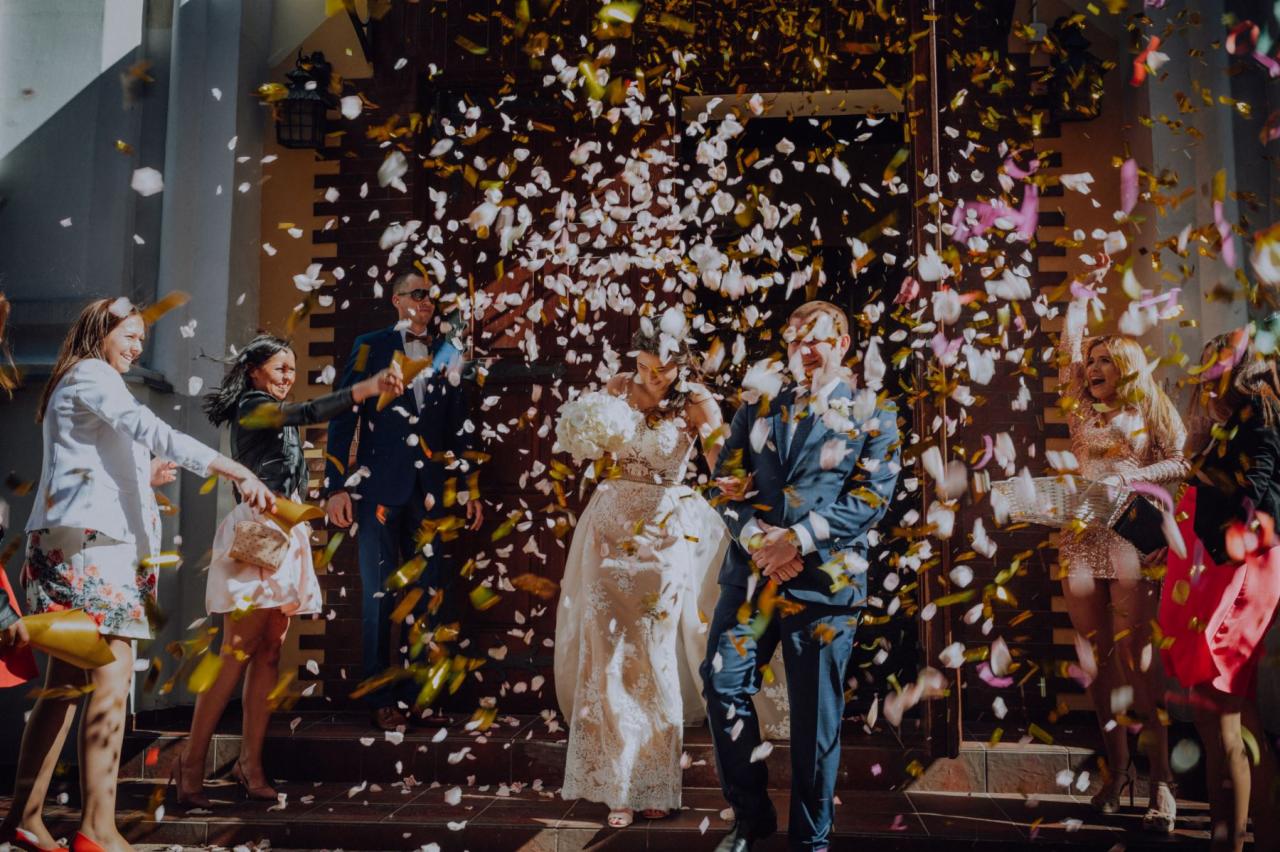Para młoda wychodzi z kościoła obsypywana konfetti przez gości, w tle drewniane drzwi i fragment ceglanej fasady budynku.