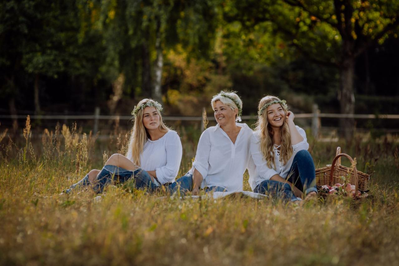 Trzy kobiety w wiankach siedzą na kocu na łące, uśmiechnięte, w tle drzewa i koszyk piknikowy.