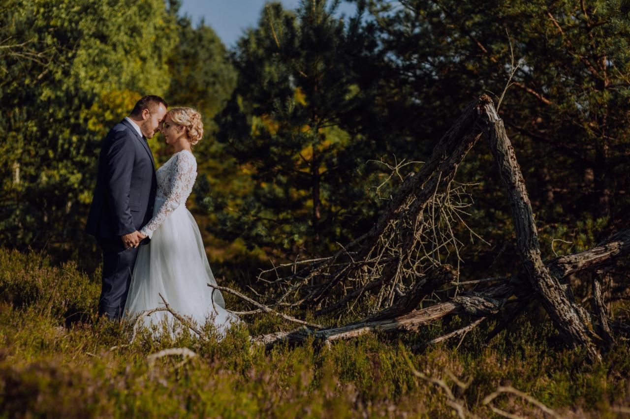 Para młoda w lesie, panna młoda w koronkowej sukni z długim rękawem i upiętymi włosami, pan młody w granatowym garniturze, stoją blisko siebie na tle drzew i powalonego pnia.