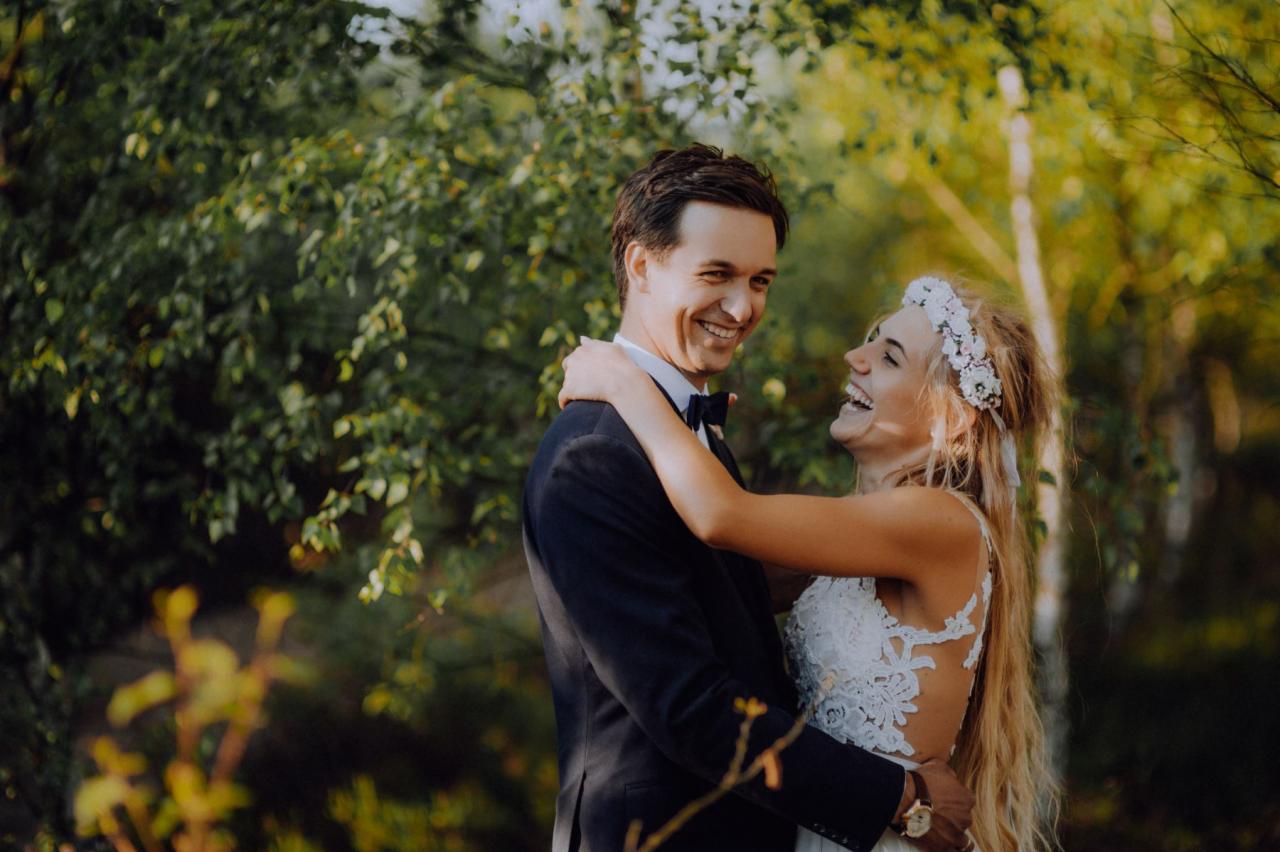 Uśmiechnięta para młoda w objęciach na tle zieleni, panna młoda z wiankiem z białych kwiatów, pan młody w ciemnym garniturze z muchą.