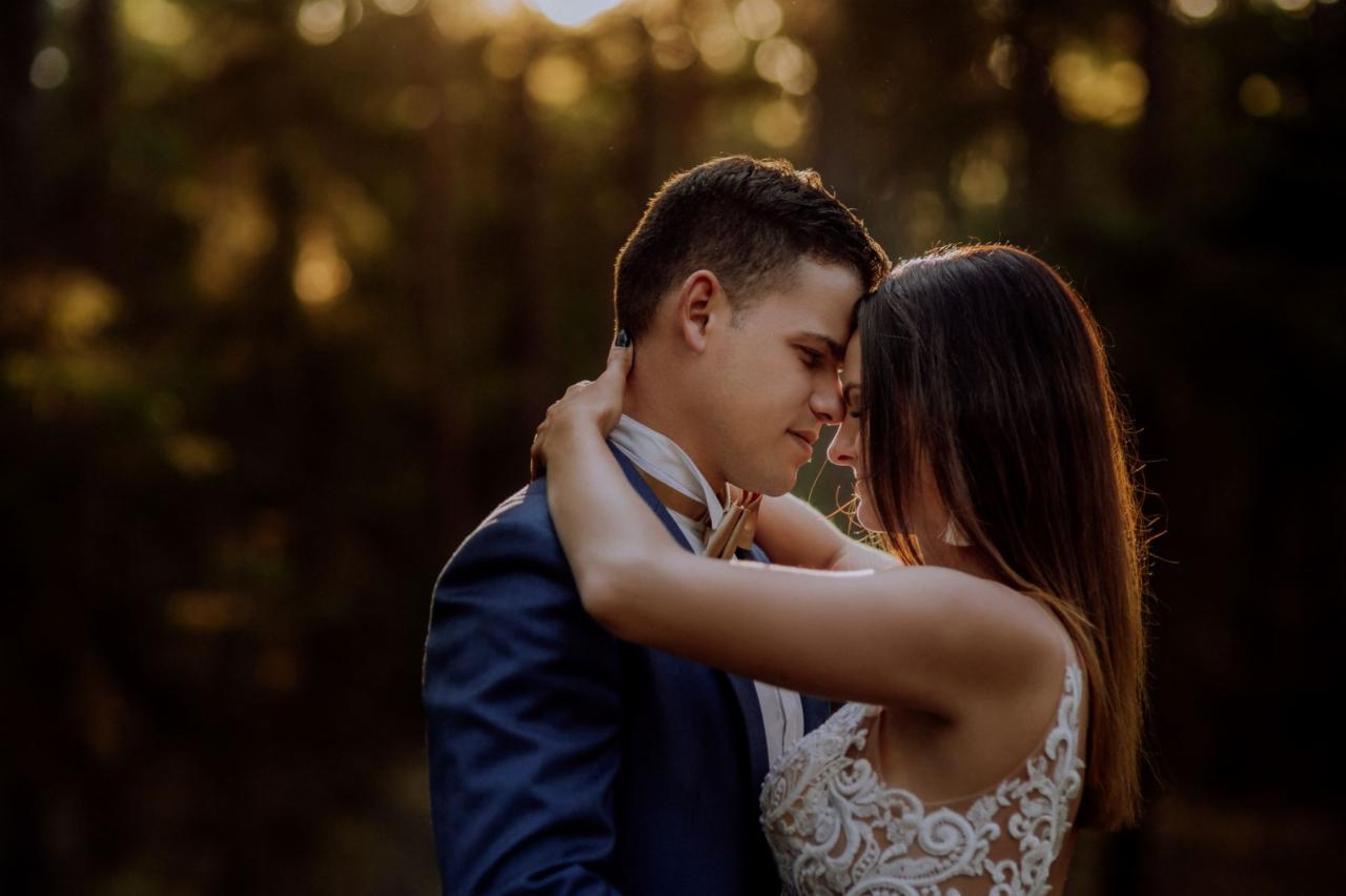 Para młoda obejmująca się czule na tle rozmytych drzew, panna młoda w koronkowej sukni, pan młody w granatowym garniturze z muszką.