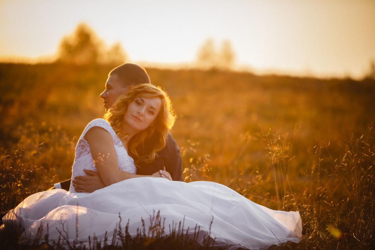 Para młoda siedząca na łące w Drawsku Pomorskim oświetlona zachodzącym słońcem, panna młoda z blond lokami spogląda w obiektyw.