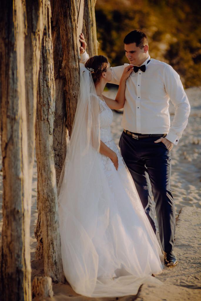 Para młoda w strojach ślubnych pozuje oparta o drewniane pale na plaży, panna młoda poprawia muchę panu młodemu, ciepłe światło zachodzącego słońca.