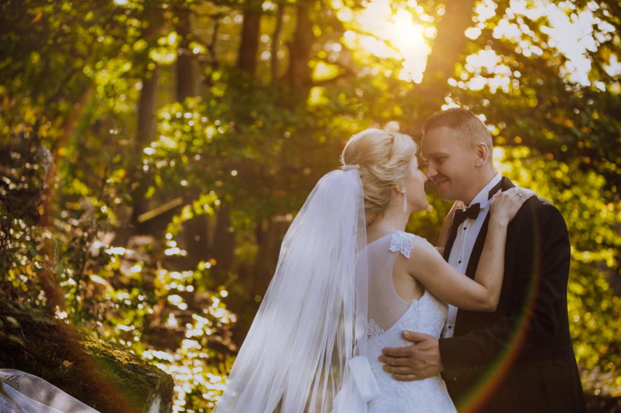 Para młoda w objęciach na tle leśnej scenerii, delikatne promienie słońca przebijają przez drzewa, tworząc ciepłą atmosferę.
