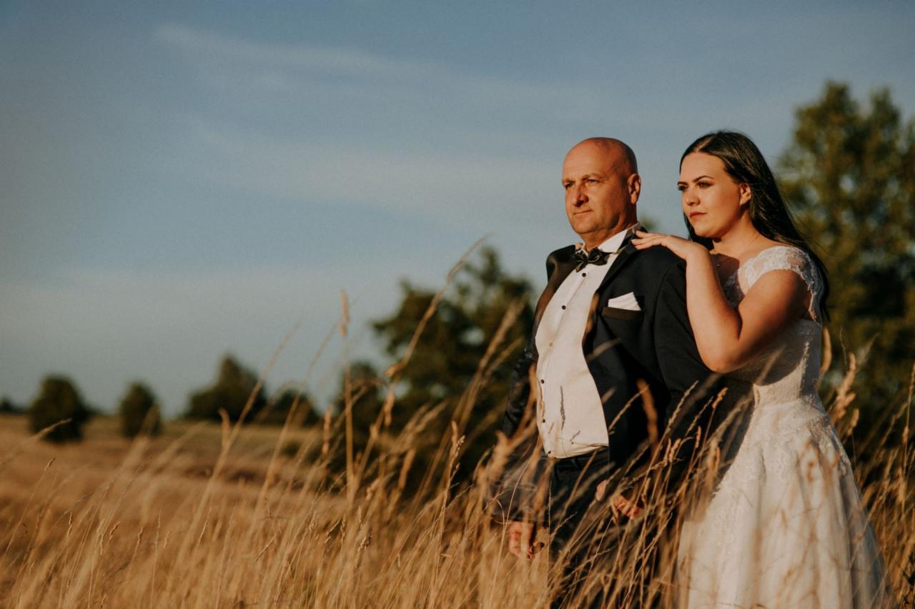 Para młoda w strojach ślubnych, pozująca na tle pola ze złotymi trawami w promieniach zachodzącego słońca.