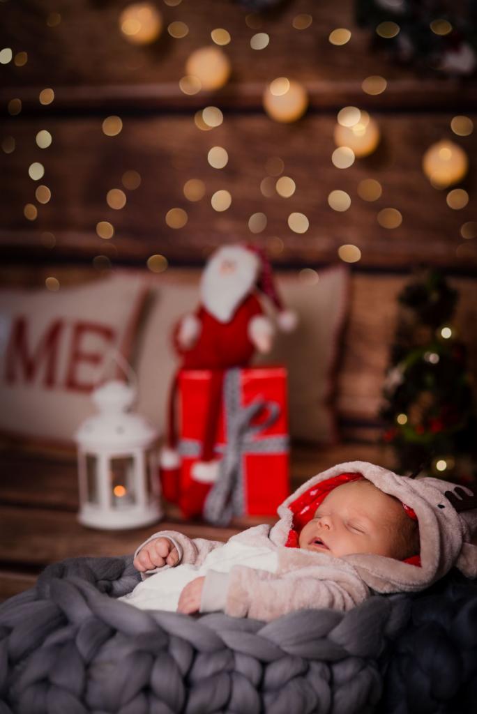 Śpiący noworodek w szarym, grubym, dzierganym koszu, ubrany w kombinezon z kapturem w kształcie renifera, na tle drewnianej ściany z lampkami, prezentem i figurką Mikołaja.