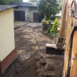 Żółta koparka w trakcie prac ziemnych na posesji z budynkami w tle, widoczne wykopane fragmenty ziemi i gruz.