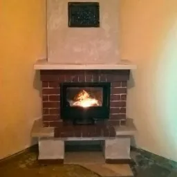 Tradycyjny kominek z czerwonej cegły z żeliwnym wkładem, płomienie widoczne przez szybę, nad nim tynkowana obudowa z ozdobną płytką, żółte ściany w tle.