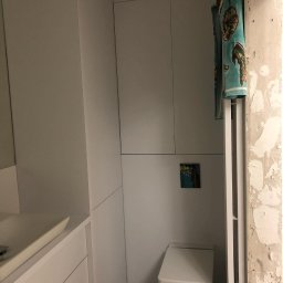 Ganes-Kuchnie i meble na wymiar Głogów - Biała zabudowa łazienkowa z szafkami, umywalką i toaletą, z widocznym fragmentem ściany w stanie surowym i suszarką na ręczniki z turkusowym ręcznikiem w morskie wzory.