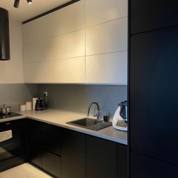 Ganes-Kuchnie i meble na wymiar Głogów - Nowoczesna kuchnia z czarnymi dolnymi szafkami i białymi górnymi, szary blat, czarny zlew i armatura, oświetlenie punktowe na suficie, biała podłoga z płytek imitujących marmur.