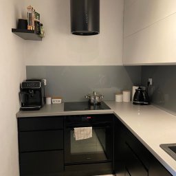 Ganes-Kuchnie i meble na wymiar Głogów - Nowoczesna kuchnia w czerni i bieli z szarym szkłem na ścianie, wyposażona w ekspres do kawy, czajnik elektryczny i płytę indukcyjną z garnkiem.