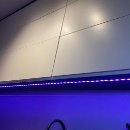 Ganes-Kuchnie i meble na wymiar Głogów - Białe, lakierowane fronty szafek kuchennych podświetlone fioletowym paskiem LED, widoczny fragment zlewu i akcesoriów kuchennych.