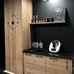 Ganes-Kuchnie i meble na wymiar Głogów - Nowoczesna kuchnia z drewnianymi frontami szafek, czarnym blatem i ścianą, dekoracyjnym zegarem ściennym i półką z ozdobami.