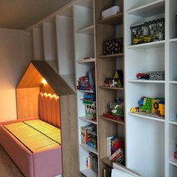 Ganes-Kuchnie i meble na wymiar Głogów - Pokój dziecięcy z zabudową meblową: różowe łóżko w kształcie domku z oświetleniem wewnętrznym, połączone z regałem na zabawki i książki, całość w odcieniach bieli, beżu i różu.
