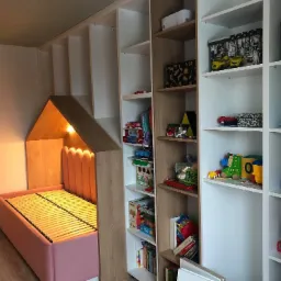 Pokój dziecięcy z zabudową meblową: różowe łóżko w kształcie domku z oświetleniem wewnętrznym, połączone z regałem na zabawki i książki, całość w odcieniach bieli, beżu i różu.