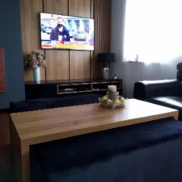 Nowoczesny salon z drewnianą ścianą telewizyjną, czarną szafką RTV, drewnianym blatem kawowym na pufie i skórzanym fotelem przy oknie.