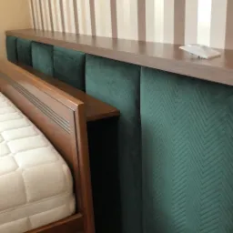 Tapicerowany zagłówek w kolorze butelkowej zieleni z geometrycznym wzorem, połączony z drewnianą półką i ramą łóżka z materacem, na tle ściany w pionowe pasy.