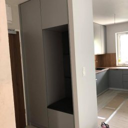 Ganes-Kuchnie i meble na wymiar Głogów - Szara zabudowa wnękowa z otwartą przestrzenią na siedzisko i półki, w tle widoczna kuchnia w podobnej kolorystyce, podłoga zabezpieczona taśmą malarską, puszki z farbą i akcesoria malarskie...