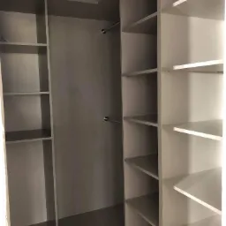 Wnętrze szafy z półkami i drążkami na ubrania, w odcieniach bieli i szarości, widok z perspektywy wnętrza pomieszczenia.