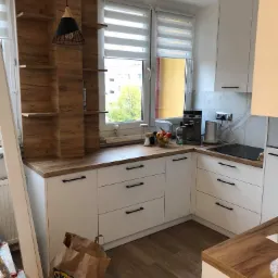 Jasna kuchnia z białymi szafkami i drewnianymi blatami, z otwartymi półkami z drewnopodobnego materiału, widoczne torby IKEA i drabina, sugerujące montaż lub wykończenie.