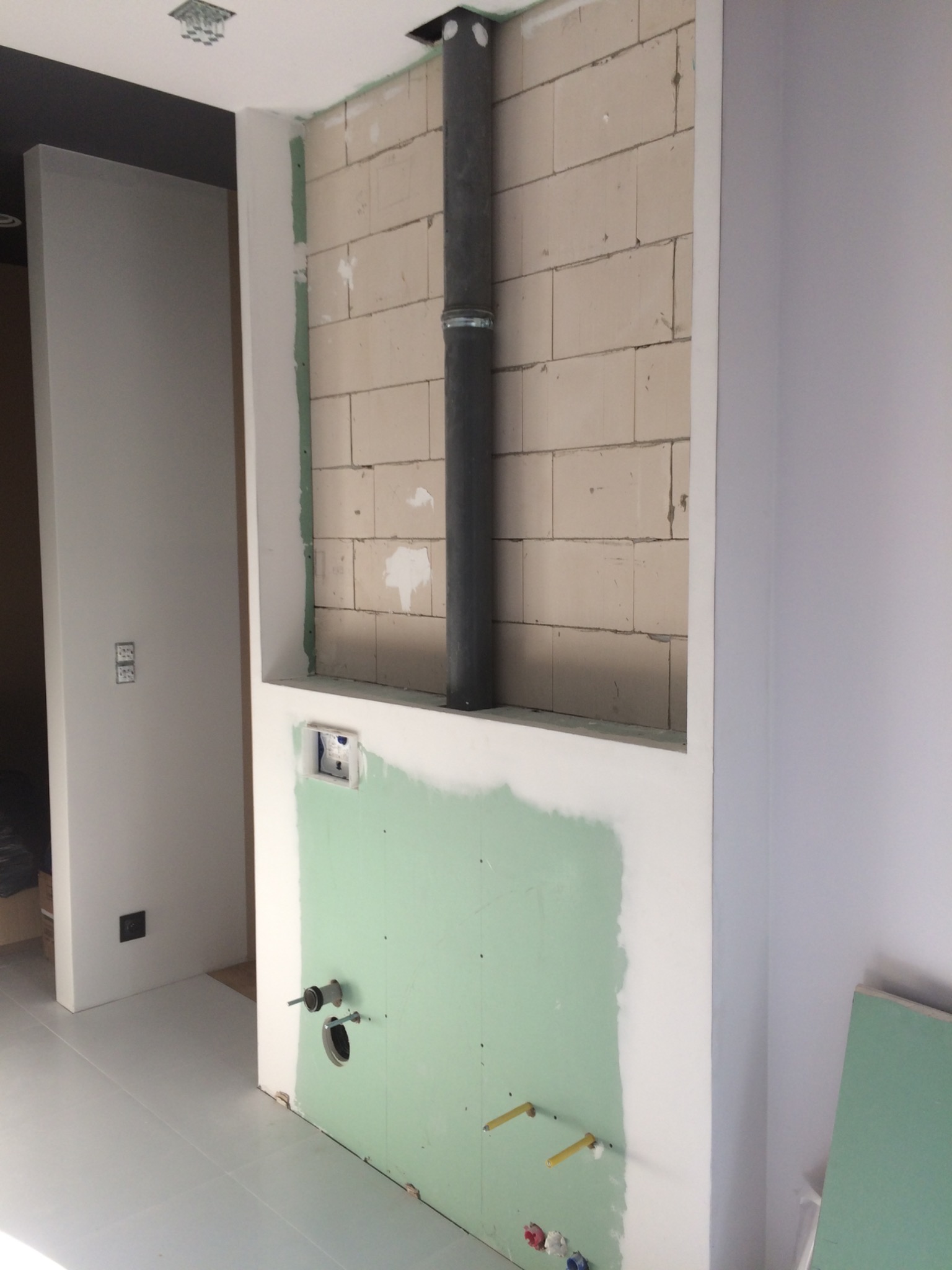 Wnęka w ścianie z surowymi blokami, pionowy przewód kominowy, instalacja wodno-kanalizacyjna w trakcie montażu, ściany częściowo pokryte zieloną płytą gipsową.