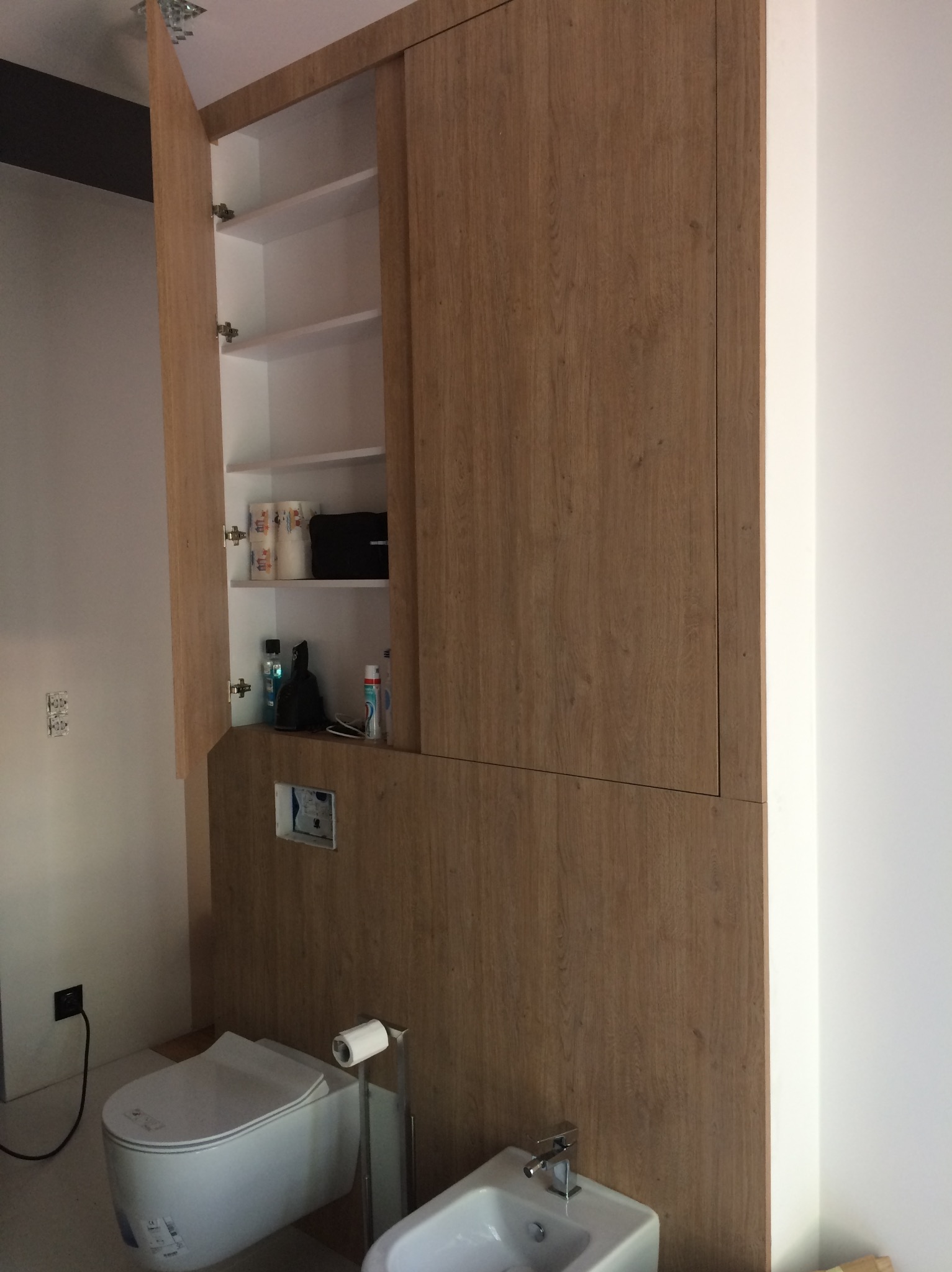Drewniana zabudowa w łazience integrująca szafę z półkami i ukrytą spłuczkę, widoczny fragment toalety i bidetu.