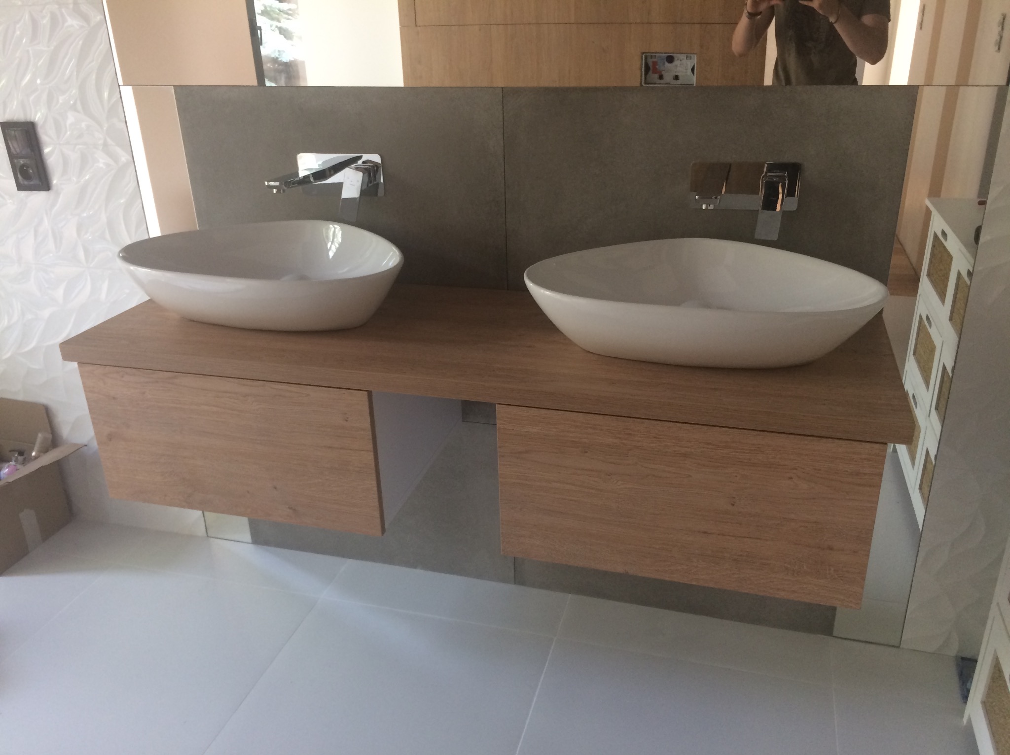 Dwie białe umywalki nablatowe z chromowanymi bateriami, osadzone na drewnianym blacie z szufladami o jasnym, dębowym wykończeniu. W tle szara ściana i lustro, w którym odbija się osoba robiąca...