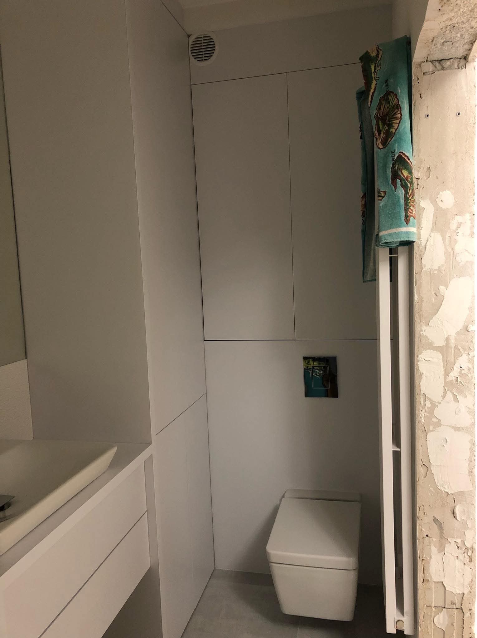Biała zabudowa łazienkowa z szafkami, umywalką i toaletą, z widocznym fragmentem ściany w stanie surowym i suszarką na ręczniki z turkusowym ręcznikiem w morskie wzory.