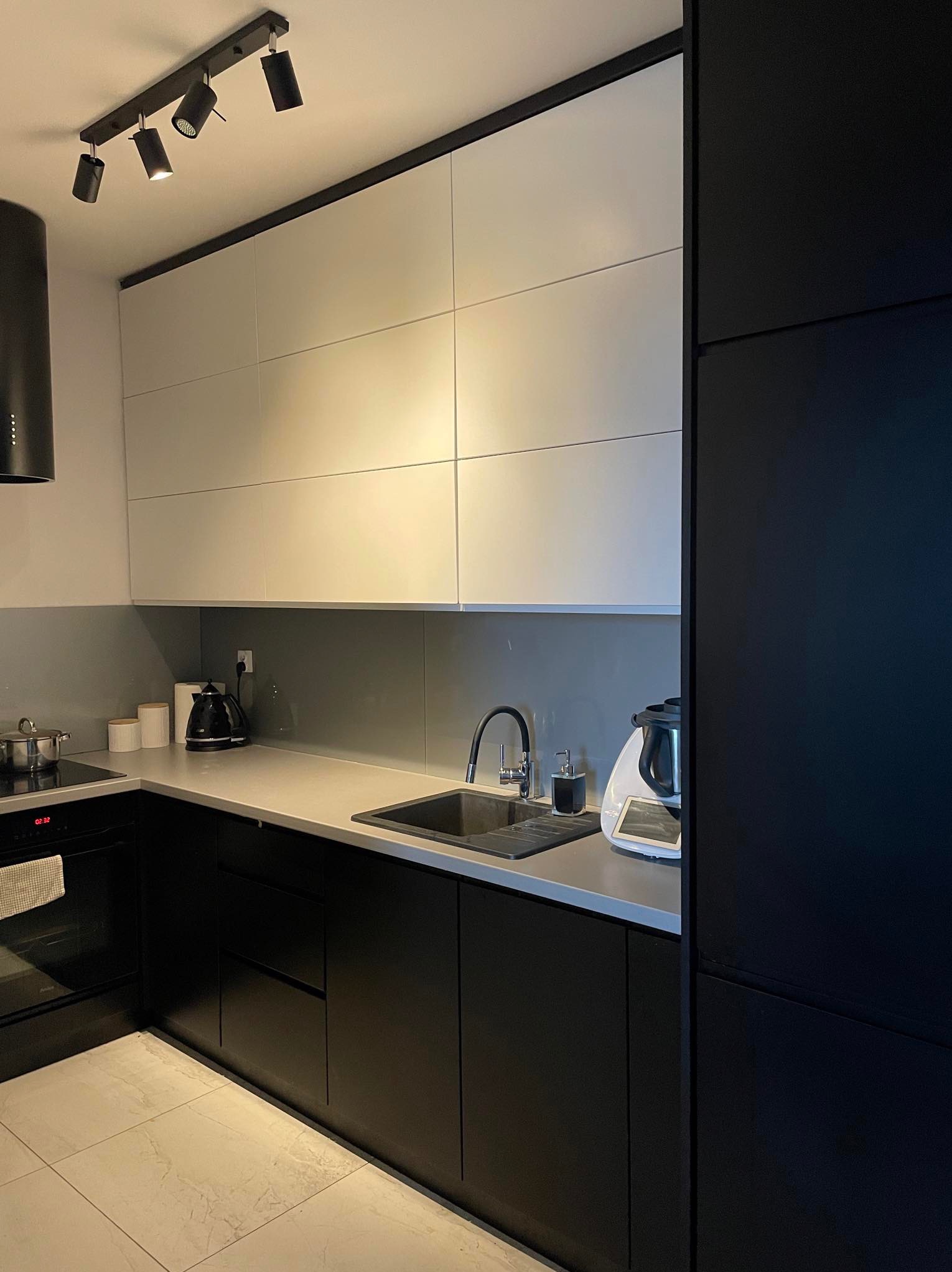 Nowoczesna kuchnia z czarnymi dolnymi szafkami i białymi górnymi, szary blat, czarny zlew i armatura, oświetlenie punktowe na suficie, biała podłoga z płytek imitujących marmur.