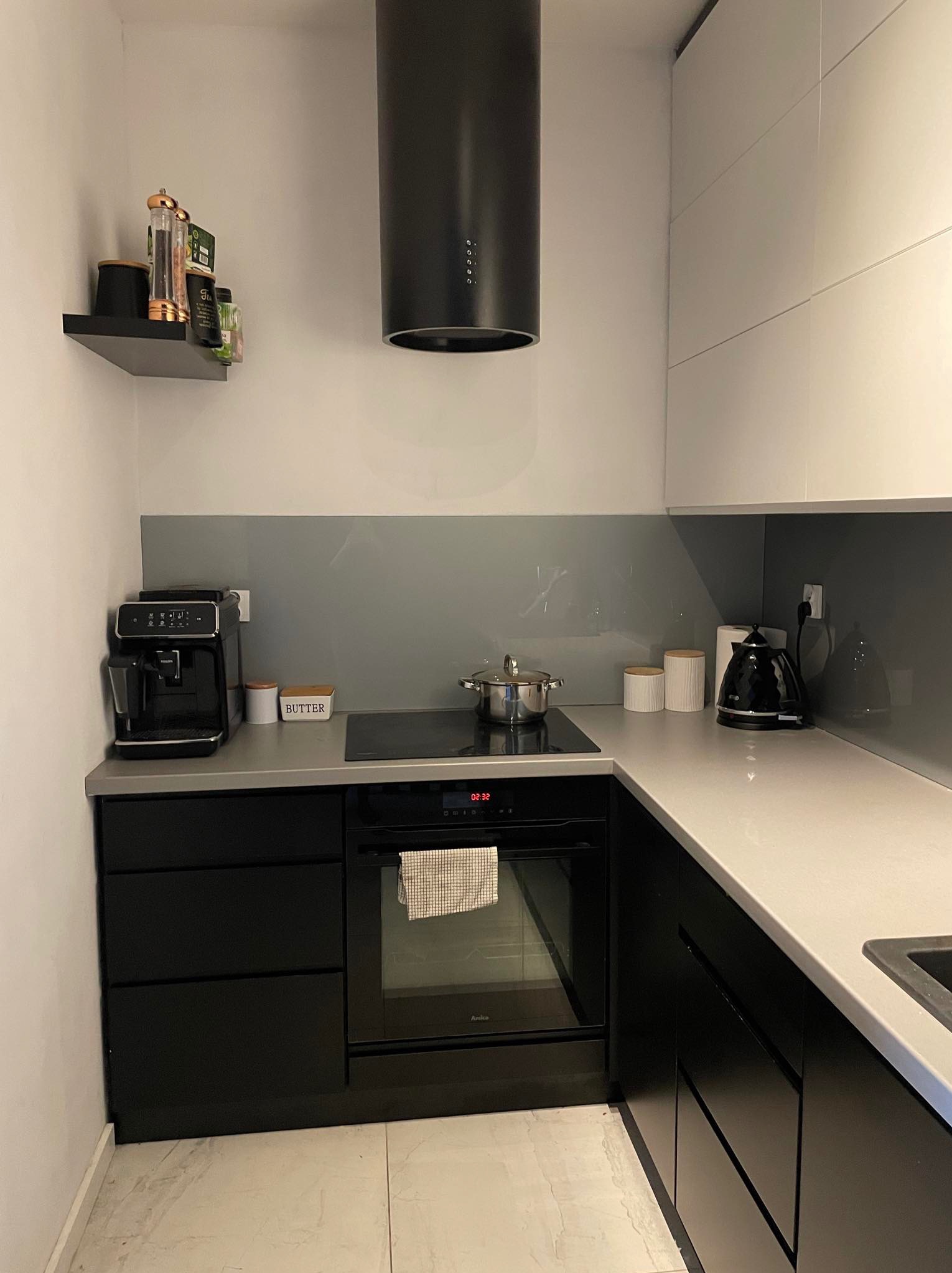 Nowoczesna kuchnia w czerni i bieli z szarym szkłem na ścianie, wyposażona w ekspres do kawy, czajnik elektryczny i płytę indukcyjną z garnkiem.