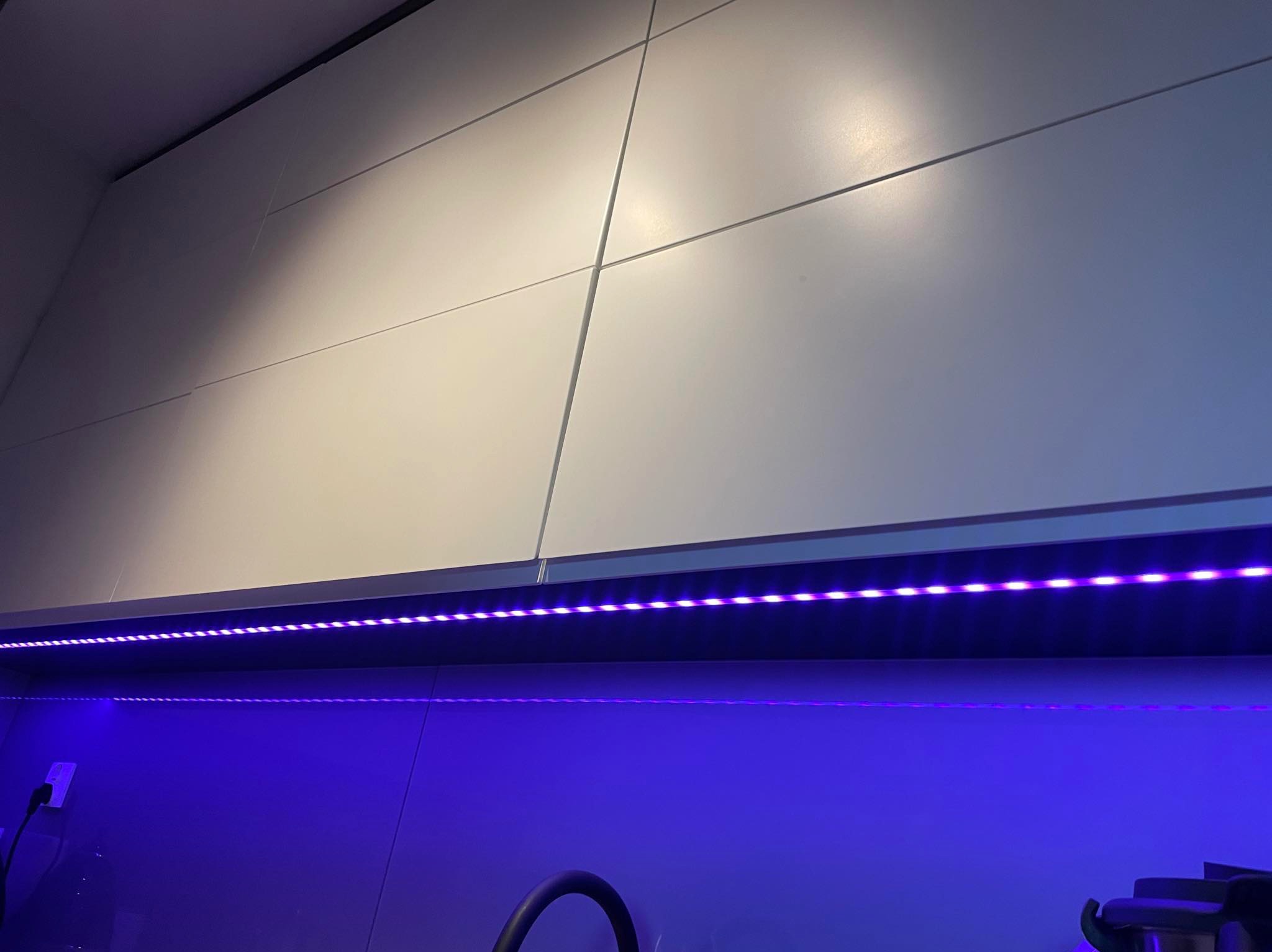 Białe, lakierowane fronty szafek kuchennych podświetlone fioletowym paskiem LED, widoczny fragment zlewu i akcesoriów kuchennych.