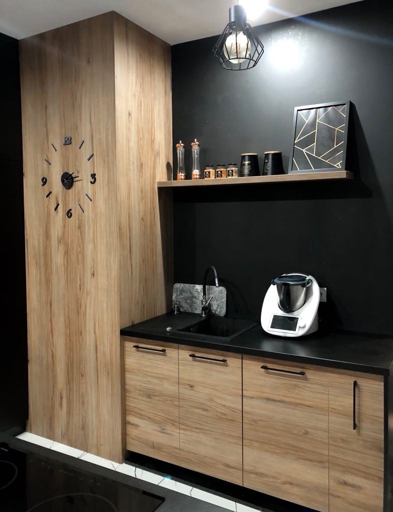 Nowoczesna kuchnia z drewnianymi frontami szafek, czarnym blatem i ścianą, dekoracyjnym zegarem ściennym i półką z ozdobami.
