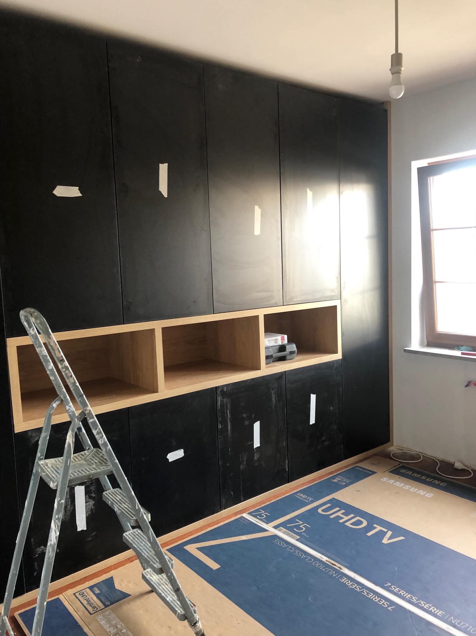 Ciemna zabudowa meblowa w trakcie montażu, z otwartymi półkami z jasnego drewna, zabezpieczona taśmą malarską, z rozłożoną drabiną i kartonami po telewizorze na podłodze.
